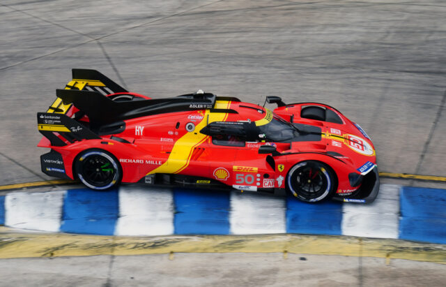 Red race car (number 50) speeding along a race track beside blue-and-white barriers, bright sponsor logos on its body.
