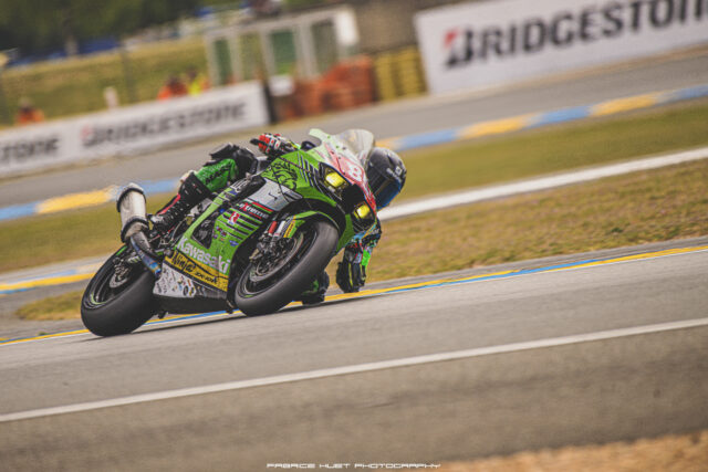 Green Kawasaki Ninja race bike leaning into a corner on a race track, rider in green and red gear with a blurred crowd and barriers in the background