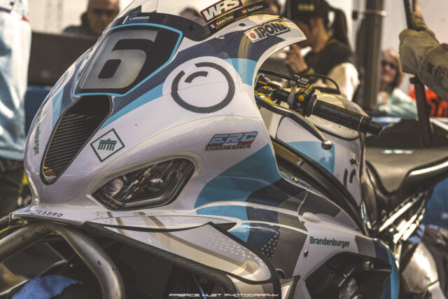 Close-up of a race motorcycle helmet with blue-white hex pattern and sponsor logos, handlebars in view, paddock behind.