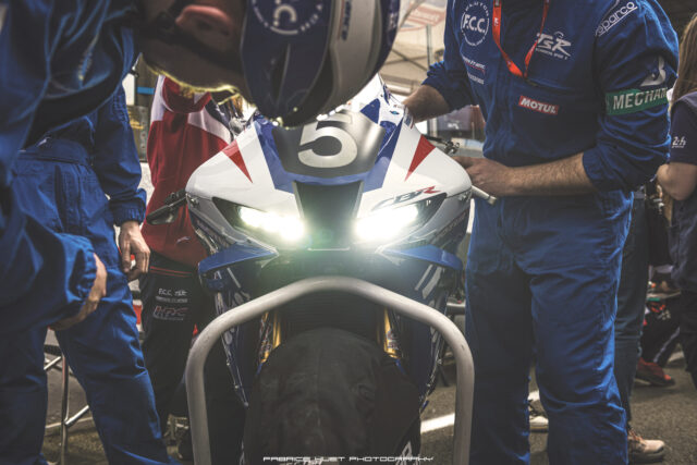 Front view of a race motorcycle with number 5, headlights on, surrounded by crew in blue pit suits during a pit stop.