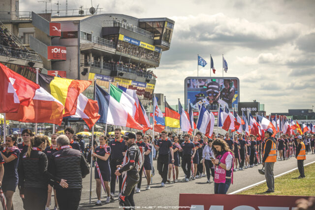 Spectators line the pit wall with numerous national flags fluttering in the foreground at a racing event.",