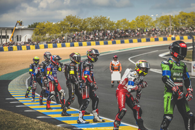 Group of motorcycle racers in full gear stand at the edge of a race track, crowds behind a blue-yellow barrier watching from the stands behind them.