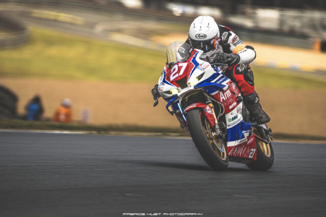 Motorcycle racer on a red, white, and blue bike leaning into a turn on a race track, number 27 visible on the bike and suit.