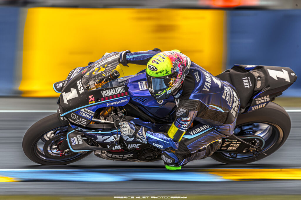 Motorcycle racer on a blue Yamaha bike leaning into a corner during a race, colorful helmet in focus.