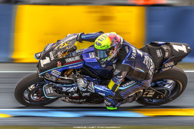 Motorcycle racer on a blue Yamaha bike leaning into a corner during a race, colorful helmet in focus.