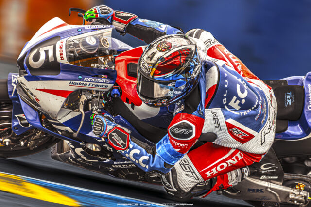 Motorcycle racer in red, white, and blue gear leaning into a turn on a race track with sponsor logos visible.