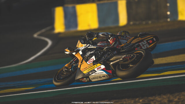 Two motorcycle racers lean into a tight turn on a sunlit race track with colorful barriers in the background, speed lines emphasized.