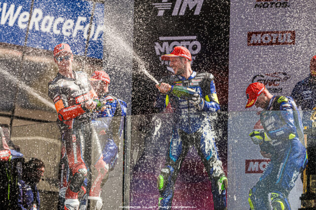 Motorcycle racers celebrate on a podium, spraying champagne amid sponsor banners like MOTUL and Yamaha.