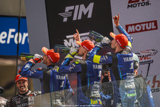 Motorcycle racers in blue Yamaha suits celebrate on a podium spraying champagne.
