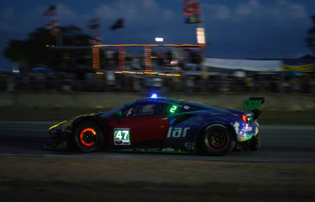 Race car #47 with blue-green livery speeds along a track at night, crowd blurred in the background.