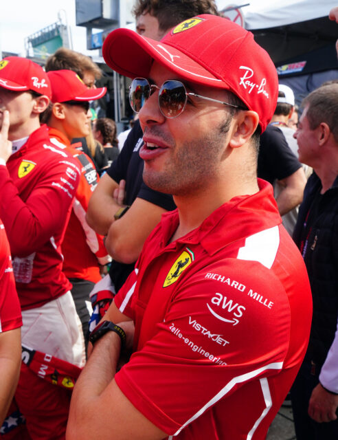 Close-up of a man in a red Ferrari cap and polo, wearing sunglasses, with sponsor logos, at a crowded motorsport event.
