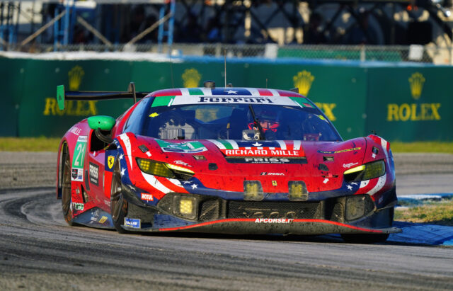 Front view of a red Ferrari race car (number 21) taking a turn on a race track, with sponsor decals visible on the hood and sides.