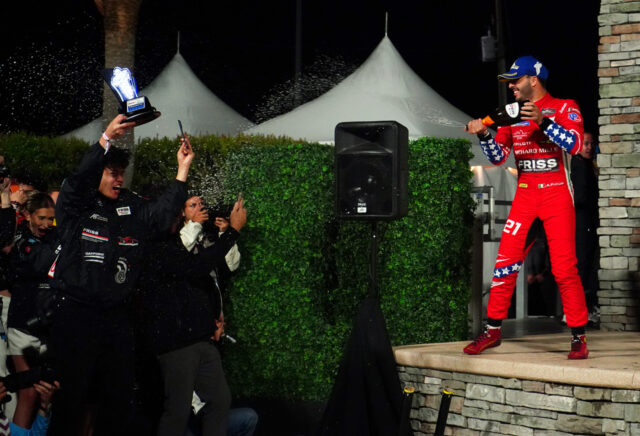 Racing driver in red suit on stage sprays champagne as crowd cheers and trophy is raised nearby at night.