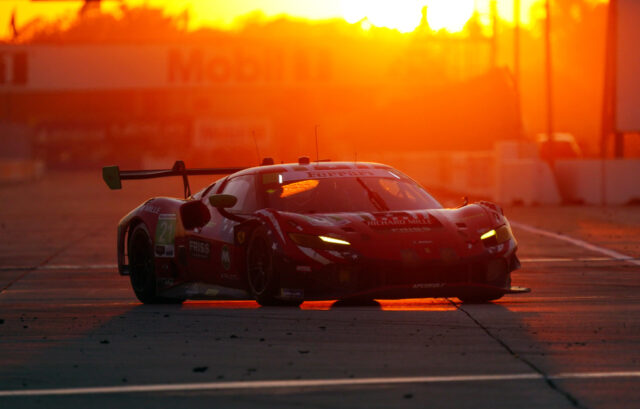 Red Ferrari racing car on a track at sunset with a bright orange sky and a large rear wing, number 21 visible on the door.
