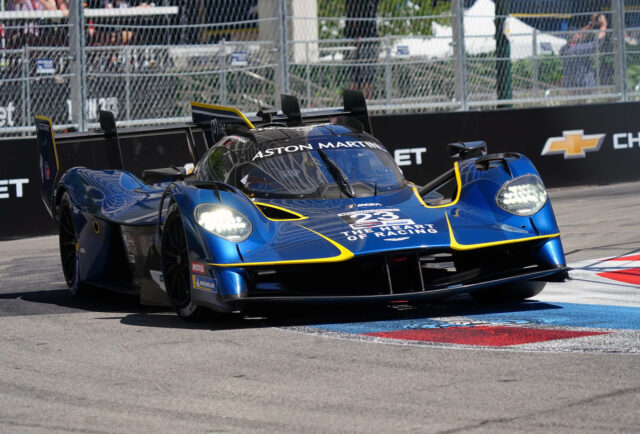 Blue Aston Martin race car (prototype) number 28, with yellow accents, speeding on track with barrier in background.