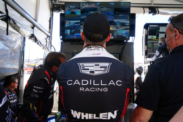 Cadillac Racing team member in a navy racing suit stands in a pit area, facing a wall of monitors showing multiple camera feeds.