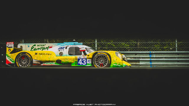 Colorful prototype race car with number 43 on a track, sponsor logos visible on its body, nighttime setting nearby railings behind fence
