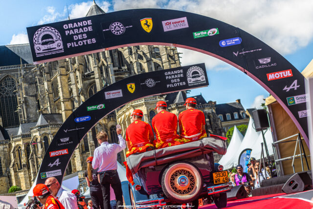 Participants in red uniforms ride a vintage car on a parade float under a sponsor arch with a historic cathedral in the background; bright day, crowd watching.