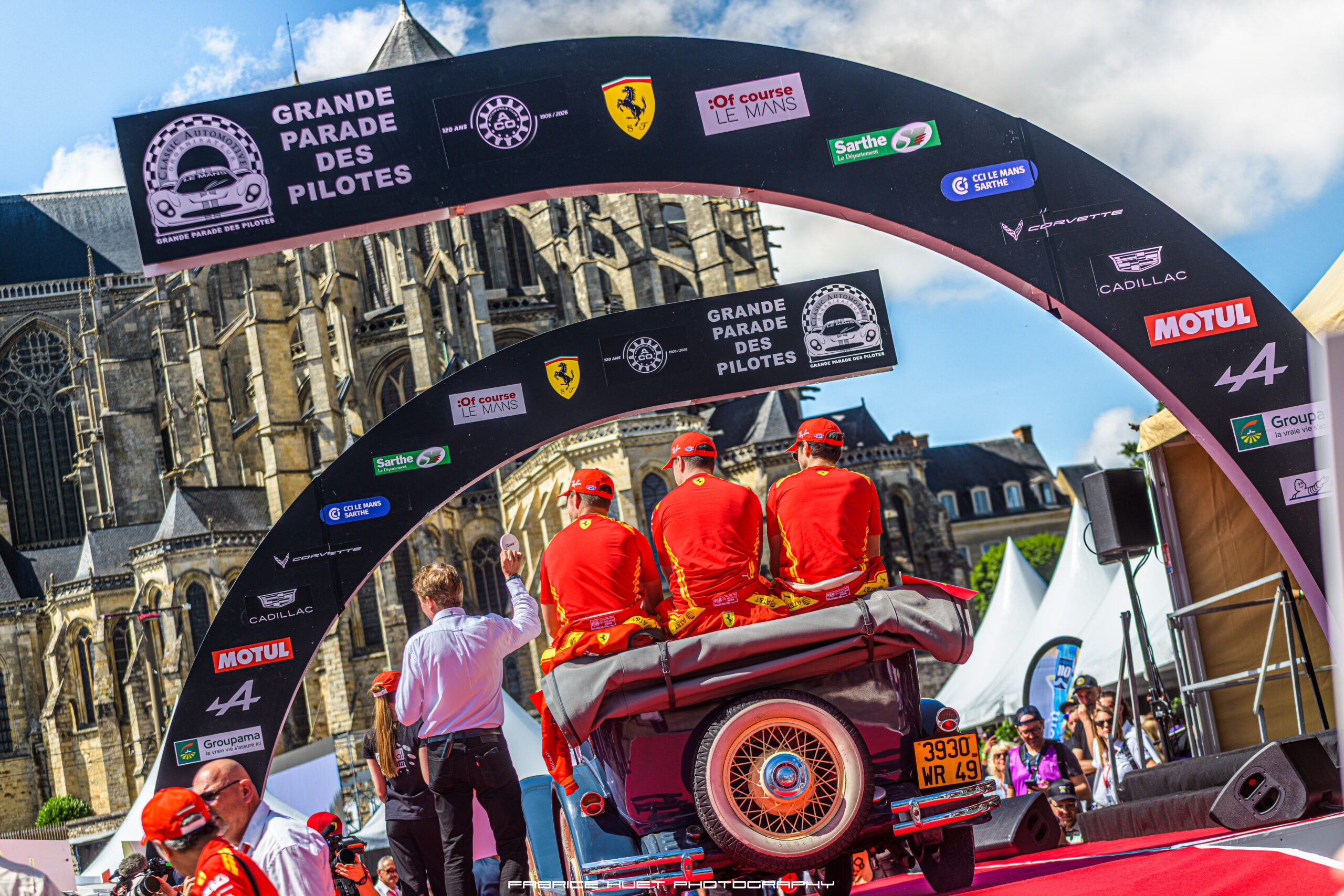 Participants in red uniforms ride a vintage car on a parade float under a sponsor arch with a historic cathedral in the background; bright day, crowd watching.