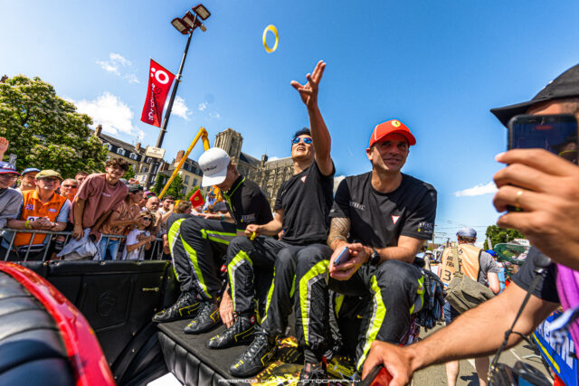 Crowd watches as athletes in black outfits toss a yellow ring into the air during a sunny street event, blue sky in the background.