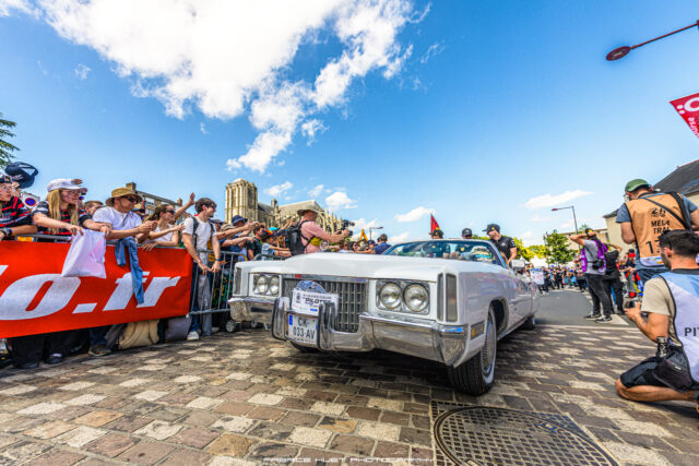 Crowd of spectators along a parade barricade watching a white vintage convertible drive past, with a Gothic church in the background under a blue sky.