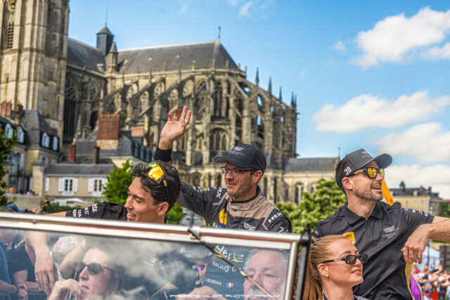 Group of race car drivers in a convertible, waving, with a Gothic cathedral and blue sky behind.