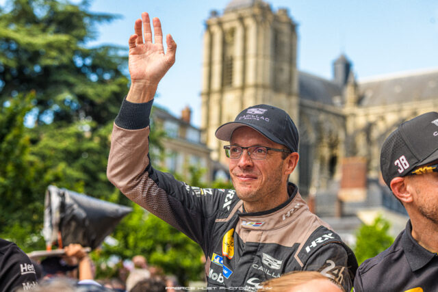 Racer in beige and black gear raises his hand in celebration, with a historic building visible in the blurred background.