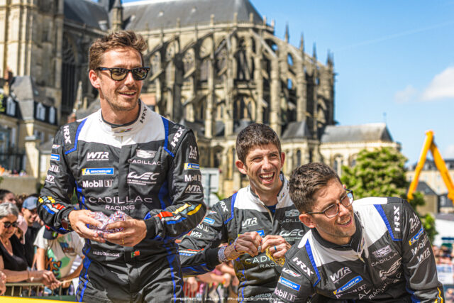 Racing drivers in Cadillac Racing suits smiling at a street event with a historic cathedral in the background and cheering crowd behind barriers.