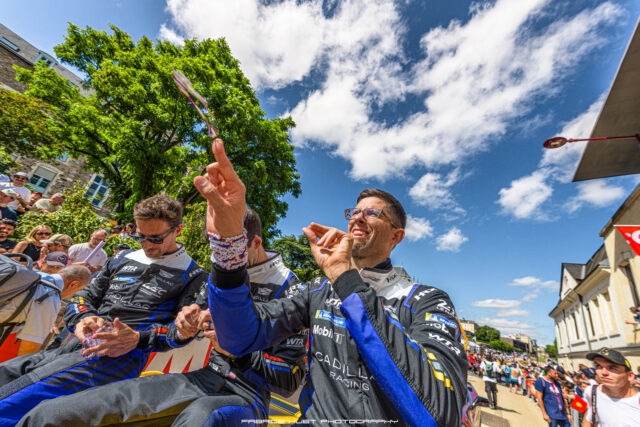 Group of rally drivers in matching racing suits celebrate on a sunlit city street, spectators watching in the background.