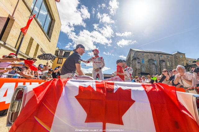 Racers on a street podium shake hands as fans cheer, with red‑and‑white banners and a bright blue sky above.