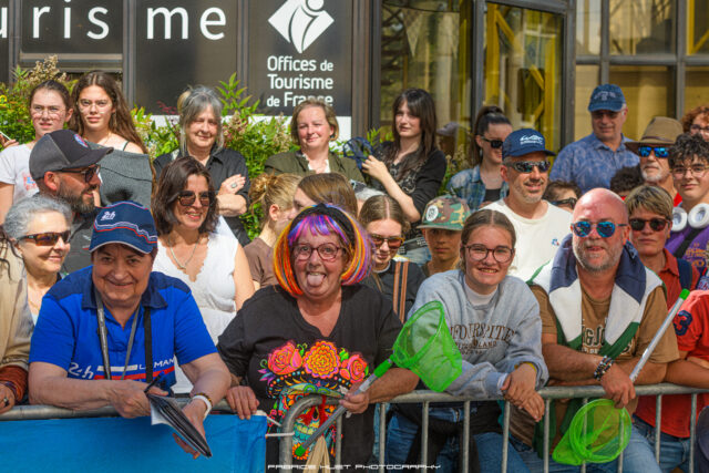 Group of diverse spectators behind a metal barrier at a public event, some smiling at the camera, with a storefront sign visible in the background.