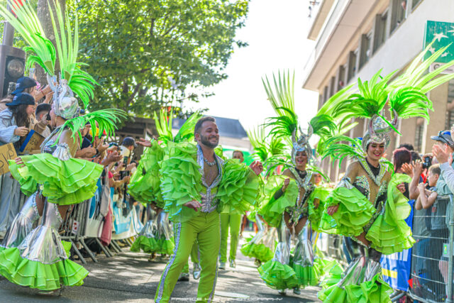 Parade dancers in lime-green costumes with tall feather headdresses perform along a crowded street.