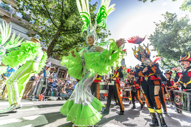 Parade participants in bright green and glittery costumes marching along a street with spectators behind barricades.