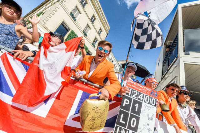 Parade participants on a float waving a Union Jack banner, smiling under a bright blue sky.