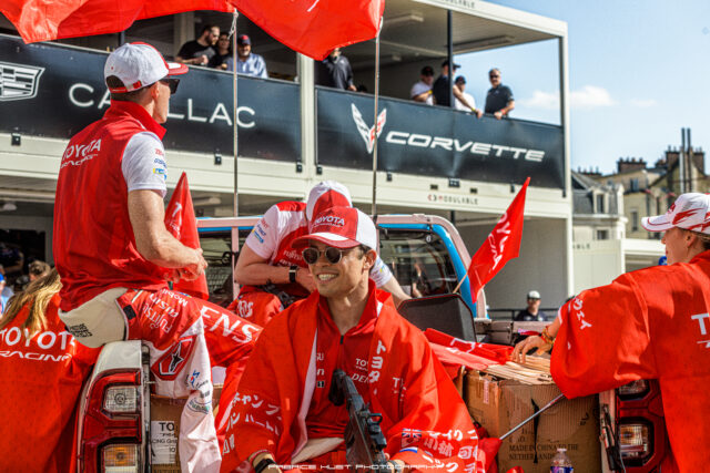 Red-clad race team members inside a pickup bed packing boxes, with a Corvette banner in the background and onlookers above in a pit-area setup.