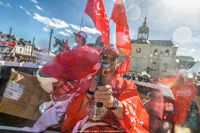 Man in red racing suit and cap leans out of a race car, holding a camera while red flags wave and spectators cheer in a sunny city square.