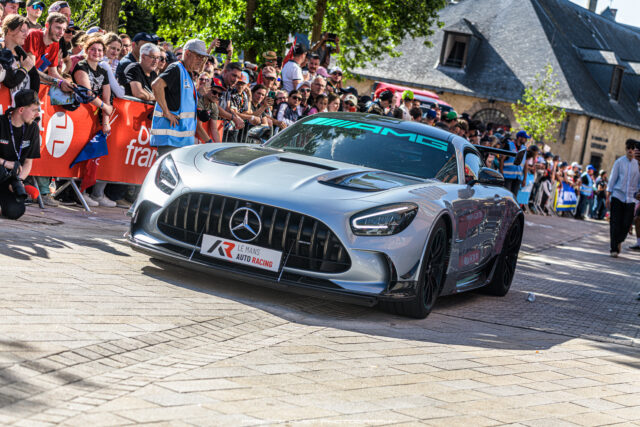 Silver Mercedes AMG race car driving past a crowded street with red barriers and cheering spectators lined along the course.