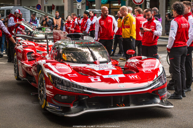 Red Toyota racing car in a pit area with a crowd of crew members in red jackets surrounding it, photographers in the background, car number 7 visible on the front bumper and sponsor decals across the body.
