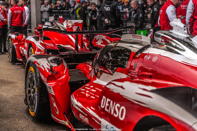 Red race cars lined up in a pit area with team members and spectators in the background behind barriers and fencing.