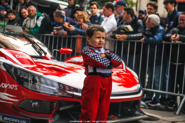 Young boy in a red and black racing suit stands with arms crossed beside a red race car as fans watch behind a barrier.