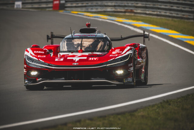 Red race car seen from the front on a racetrack, with a large rear wing and sponsor decals visible on the bodywork.