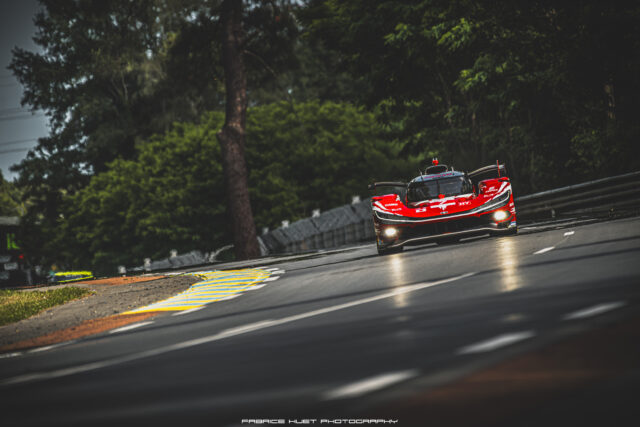 Red race car on a curving outdoor track, surrounded by green trees, speeding toward the viewer with headlights on and sponsor logos visible on the body.