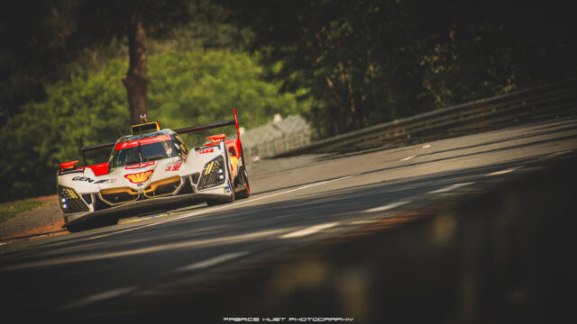White and red GT race car takes a left-hand turn on a tree-lined race track, sponsor logos visible across the nose and cockpit area.