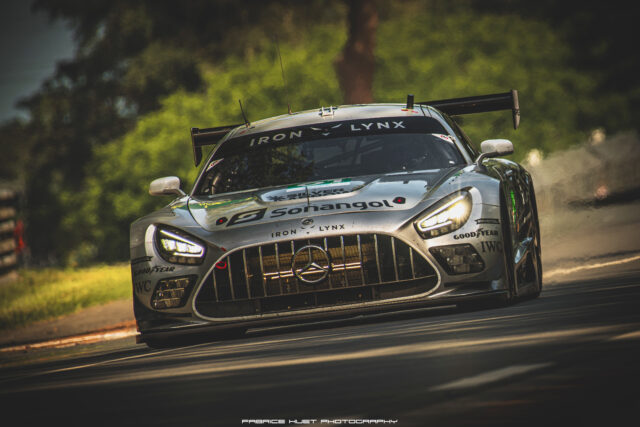 Silver race car speeding through a turn, front view with sponsor logos and prominent grille (Iron Lynx). A high-speed motorsport image on a race track, rear wing visible.