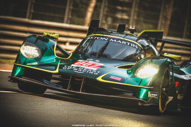 Green Aston Martin racing prototype on a track, front view with headlights on and sponsor logos visible.