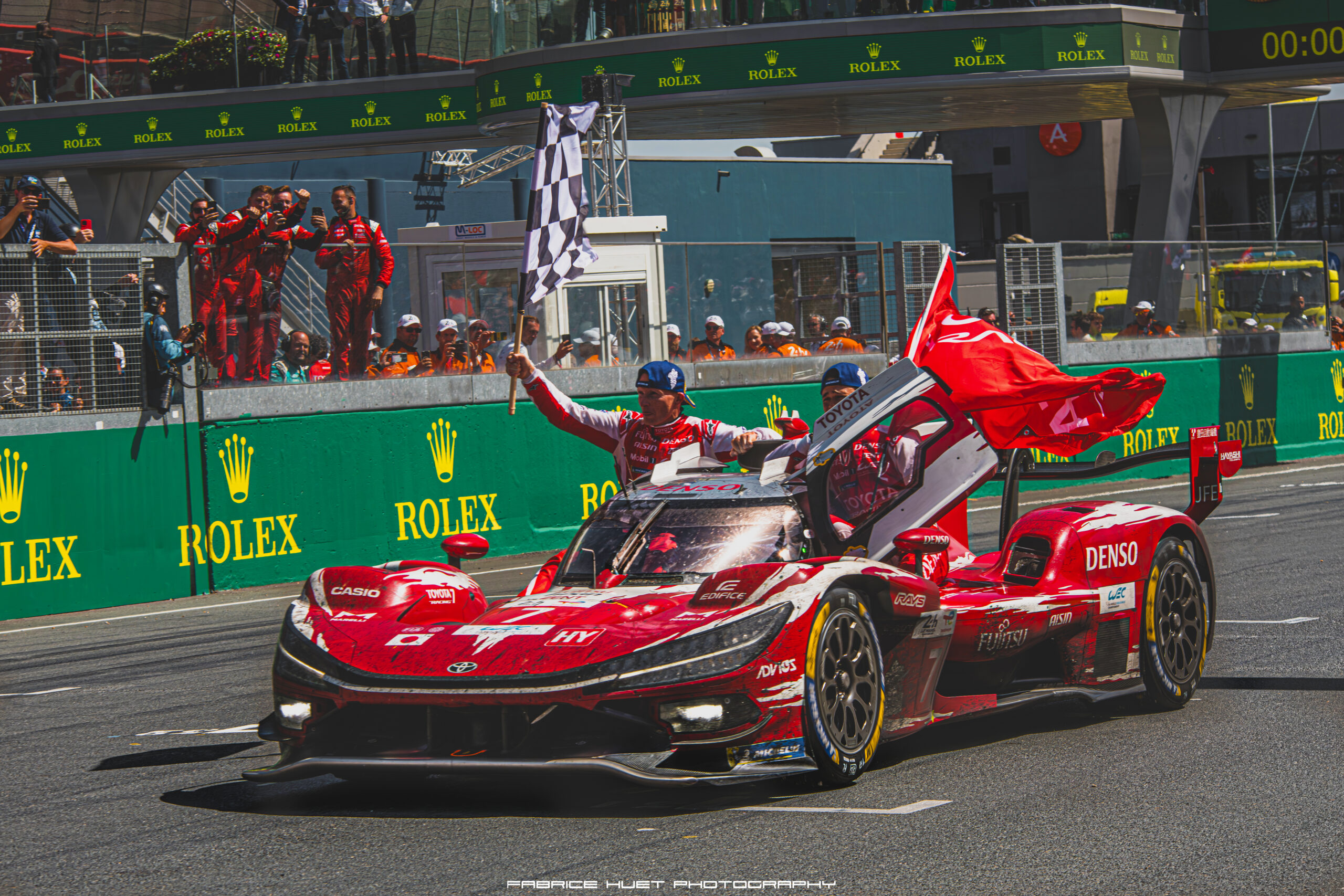 Red race cars pull into the pit wall as photographers and crew cheer from the pit lane, checkered flag raised for the win against a Rolex sponsor backdrop.