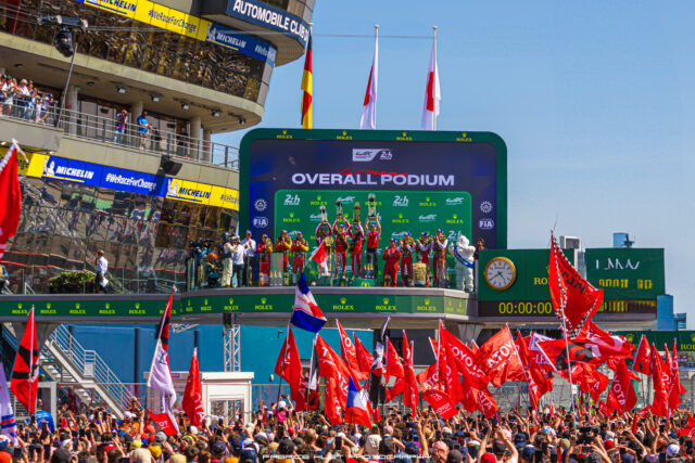 Victory podium celebration at a motorsport race with drivers raising trophies, a large LED screen behind showing podium text, and a cheering crowd with red flags.