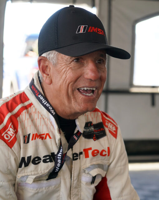 Older man in a white and red WeatherTech racing suit, wearing a black IMSA cap, smiling in a pit area.