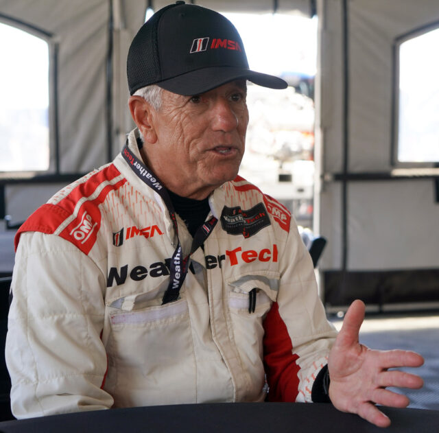 Man in a white-red racing suit with WeatherTech and IMSA logos, wearing a black cap, speaking and gesturing with his hand.