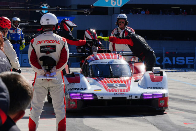Pit crew works on a white-red Porsche race car in the pit lane, one member wearing an 'OFFICIAL' suit and others nearby helping during a pit stop at speedway.